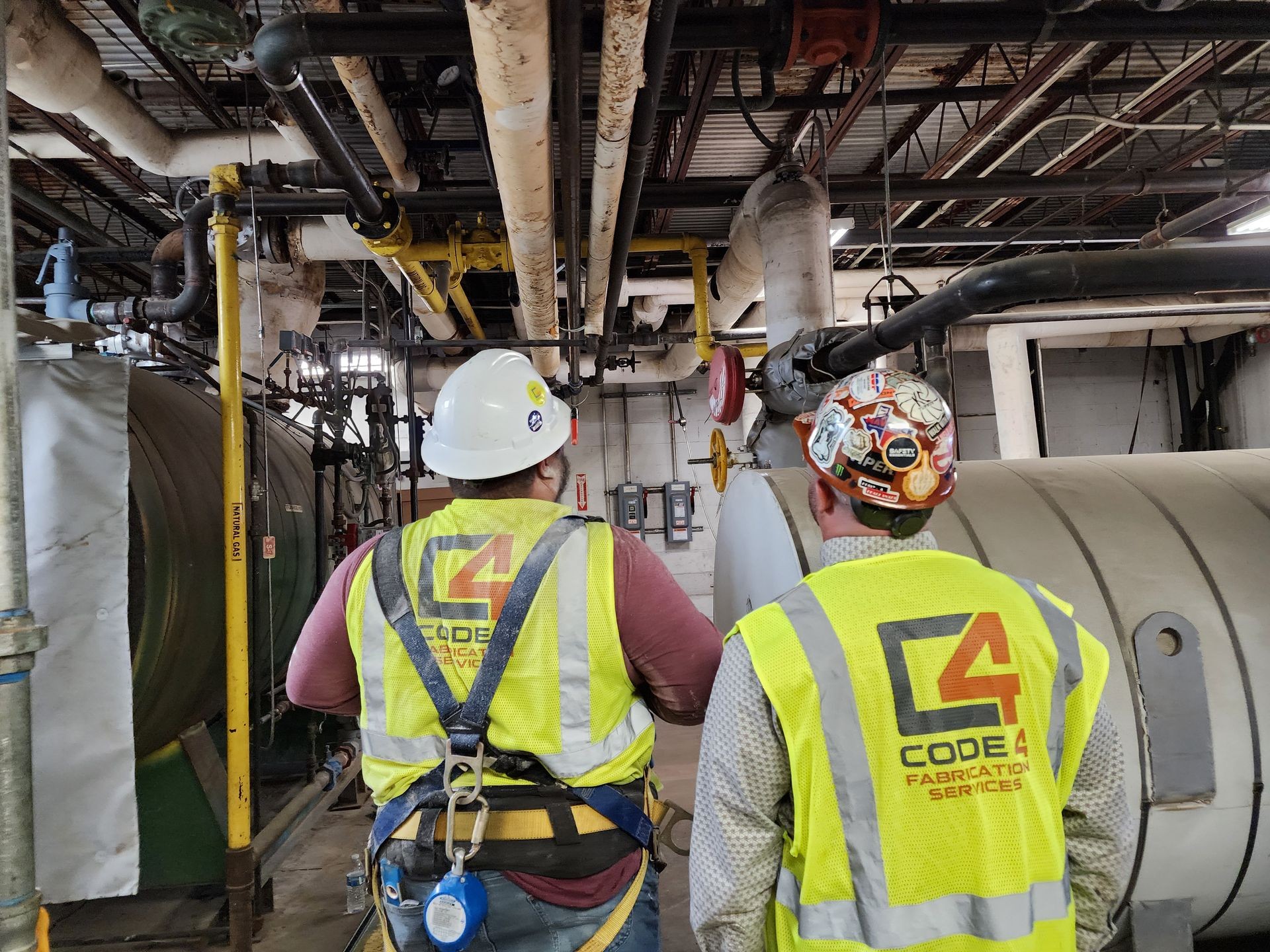 Two workers in safety vests and helmets inspecting industrial pipes and machinery in a factory setting.